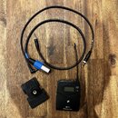 Na dřevěné podlaze je rozložen bezdrátový vysílač značky SENNHEISER s označením ew G4, černý plastový držák a černý kabel. Kabel má na koncích jack k…