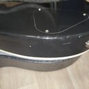 Detail zadní strany černé elektrické kytary s bílým lemováním. Je vidět krycí deska elektroniky připevněná třemi šrouby a kovová zásuvka pro kabel. L…