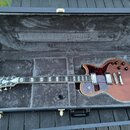 Elektrická kytara s hnědým trupem a tmavě červeným pickguardem v černém tvrdém kufru. Na hlavě hmatníku je bílé logo "Epiphone". Nástroj má dva humbu…