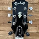 Černá hlava elektrické kytary s bílým logem Epiphone v horní části a bílou stylistickou ikonou uprostřed. Na zadní straně hmatníku je viditelný nápis…