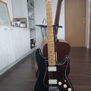 Černá elektrická kytara s červeně vzorovaným pickguardem a světle hnědým hmatníkem. Na hlavě nástroje je viditelné logo Fender. Kytara stojí na stoja…