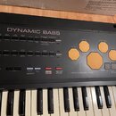 Černý elektronický klávesový nástroj s bílými a černými klávesami. Na horním panelu je nápis DYNAMIC BASS a sekce PERCUSSION PAD s oranžovými polštář…