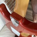 Detail zadní strany krku a těla elektrické kytary v červené barvě. Krk je vyroben ze světlého dřeva s lakovaným povrchem. Na spojení krku s tělem je…