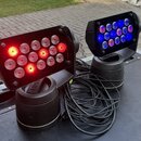 Dvě černé LED par lampy s pohyblivým základnami, umístěné na černém transportním kufru. Levá lampa svítí červeně a žlutě, pravá modře a fialově. Na h…