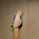 Fotografie zachycuje hlavu a horní část hmatníku elektrické kytary z přírodního dřeva. Na hlavě je vidět logo Fender a nápis STRATOCASTER®. Nástroj j…