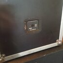 Zadní strana černé reproduktorové bedny v hliníkovém transportním kufru. Uprostřed je umístěn ovládací panel s nápisem MUSICAL INSTRUMENT SPEAKER ENC…