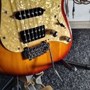 Detail elektrické kytary s tělem v barevném přechodu od žluté po červenou. Nástroj má perleťový pickguard, dva černé snímače a černý tremolo systém s…