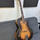 Kytara typu archtop s tmavě hnědým sunburst povrchem a bílým lemováním. Na hlavě jsou vidět čtyři ladičky a dva červené dekorativní prvky. Tělo má dv…