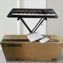 Černý digitální klavír umístěný na černém X-stojanu. Pod nástrojem je hnědý kartonový obal s nápisy YAMAHA a PSR-SX600. Na krabici leží plastový obal…
