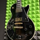 Černá elektrická kytara s tvarem single-cut a lesklým povrchem. Nástroj má dva humbuckery, čtyři ovládací knofíky a zlatý hardware. Na těle není vidě…