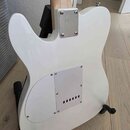 Zadní strana bílé elektrické kytary s tvarem typu Telecaster, umístěná na černém stojanu. Na zadní straně těla je bílý plastový kryt elektroniky přip…