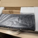 V otevřené kartonové krabici s polystyrenovými výplněmi je černý předmět obalený v průhledné plastové fólii. Vedle něj se nachází menší hnědý karton…