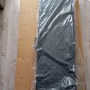 Černý obdélníkový předmět, pravděpodobně podložka nebo pouzdro, zabalený v průhledné plastové fólii. Celý předmět leží na hnědém kartonu. Na fotce ne…