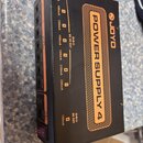 Černý obdélníkový zdroj napájení s nápisem POWER SUPPLY 4 a logem JOYO. Na horní a boční straně jsou viditelné výstupy s označením DC 9V OUT a DC OUT…