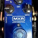 Modře třpytivý efekt pedál s nápisy MXR a BASSINNOVATIONS. Na horní části jsou tři černé ovládací knoflíky s popisky MID+, DRY, GROWL a GIRTH. Ve spo…