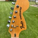 Záběr na hlavu dřevěné elektrokytary v přirozené barvě. Na povrchu je černý nápis Fender STRATOCASTER a menší text MADE IN USA. Pod logem je uveden s…