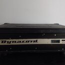 Černý zesilovač v transportním flightcase s kovovými hranami. Na přední bílé liště je nápis Dynacord a přepínač s označením ON STANDBY. Zařízení vyka…