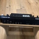 Zadní strana černého syntetizátoru s nápisy Elektron a Analog Four MKII. Zařízení je umístěno na světlém dřevěném podstavci a obsahuje řadu konektorů…