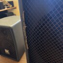 Detailní pohled na dva černé reproduktory s kovovými mřížkami. Na levém, menším reproduktoru je viditelné bílé logo HK. Pravý reproduktor vykazuje vý…