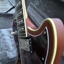 Detailní pohled na hnědou elektrickou kytaru s krémovým lemováním a tmavým hmatníkem. Nástroj má tmavě červený vzorovaný pickguard a jeden viditelný…