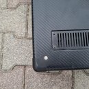Detailní pohled na černý povrch s texturou imitující karbon. Na povrchu je přišroubovaná černá ventilační mřížka s vertikálními sloty. V levém dolním…