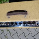 Kytarový kombozesilovač s béžovým texturovaným potahem a hnědou plastovou rukojetí s nápisem Fender. Horní ovládací panel je z chromovaného kovu a ob…