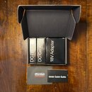 Otevřená černá krabice obsahuje tři černé obdélníkové krabičky s bílými nápisy „DC Brick Power Supply“, „DC Cables“ a „18V Adapter“. Před krabicí lež…