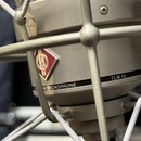 Detailní záběr na stříbrný kondenzátorový mikrofon v popfiltru. Na těle je viditelné červené kosočtvrhé logo s nápisem NEUMANN a vedle něj text N MIC…