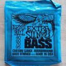 Modré plastové balení strun pro basovou kytaru. Na obalu je černý nápis ERNIE BALL EXTRA SLINKY BASS s obrázkem orla a uvedenými kalibry 40, 60, 70,…