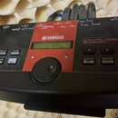 Černý modul elektronických bubnů s červeným středovým panelem. Na zařízení jsou nápisy YAMAHA a DTXPLORER DRUM TRIGGER MODULE. Modul obsahuje malý di…