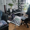 Na chromovaném stojanu je umístěn bílo-černý sampler s displejem a zelenými podsvícenými pady. Na levé straně přístroje je vidět červené logo AKAI. V…