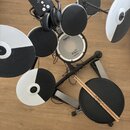 Elektronická bicí souprava s černými pady a bílo-černými činely na černém stojanu. Na modulu, snare padu a činelech je vidět nápis Roland. Na bubenká…