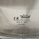 Detail bílého bubnového koženu s černými nápisy. Je zde vidět logo REMO pod stylizovanou korunkou, text DRUM HEAD a vedle něj zkratka UK. Dole je uve…