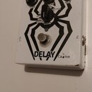 Bílý efekt pedál s černým grafickým motivem pavouka. Na horní straně jsou tři černé ovládací knoflíky s popisky TIME, REPEAT a MIX a jeden menší knof…