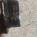 Detailní záběr na černý kovový prvek, pravděpodobně součástku hudebního nástroje. Je na něm vidět bílý nápis Ibanez a pod ním menší text LO-TRS TREM.…