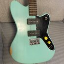 Světle tyrkysová elektrická kytara s černým pickguardem a dvěma černými humbuckery. Na hmatníku je v horní části viditelné bílé logo ve tvaru ptáka.…