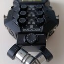 Černý přenosný záznamový systém s logem ZOOM. Zařízení má několik ovládacích knoflíků, tlačítka a malý displej v horní části. V přední části jsou umí…