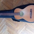 Kytara s nylonovými strunami v přirozené dřevěné barvě, položená na černém obalu. V otvoru rezonanční desky je vidět štítek s nečitelným textem. Nást…