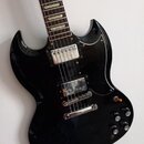 Černá elektrická kytara s typickým SG tvarem korpusu a tmavým hmatníkem s trapezoidními vložkami. Nástroj má dva chromované humbuckery, Tune-o-matic…