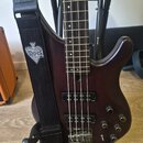 Tmavě hnědá elektrická basová kytara s čtyřstrunnovým hmatníkem a dvěma snímači. Na černém popruhu je bílo-černý nápis ACE OF SPADES v srdci. Tělo ná…