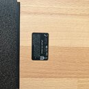 Detail spodní strany dřevěného přístroje světlé barvy s viditnou strukturou dřeva. Na povrchu je nalepený černý štítek s bílým textem a logy. Jsou na…