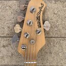 Hlava pětstrunné basové kytary z přírodního dřeva. Na povrchu je černý nápis ERNIE BALL MUSICMAN StingRay5 s doprovodnou grafikou. Jsou vidět pět kov…
