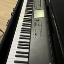 Černý digitální klavír v černém transportním kufru. Na pravé straně je vidět nápis KRONOS a pod ním menší text MUSIC WORKSTATION. Nástroj má displej,…