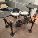 Elektronická bicí souprava na černém kovovém rámu s gumovými pady a činely. Na černém modulu je viditelné logo ROLAND. Součástí je černý kulatý sedák…