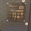 Fotografie detailu kovového štítku na černém texturovaném povrchu kytarového komba. Štítek nese nápisy Bogner AMPLIFICATION, CUSTOM SHOP a webovou ad…