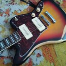 Elektrická kytara s korpusem v barevném přechodu od červené přes žlutou po světle hnědou. Nástroj má tmavě červený pickguard s texturou, dva bílé sní…