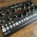 Černý syntezátor s nápisem BASSLINE a označením DB-01 v levém dolním rohu. Na pravé straně je nápis erico synthe. Přístroj obsahuje několik černých o…