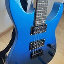 Modrá elektrická kytara s kovovým leskem a černým hardwarem. Na obou snímačích je viditelný nápis "Schecter". Nástroj je umístěn na černém stojanu a…