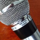 Detailní pohled na horní část dynamického mikrofonu s kovovou síťovkou a stříbrným prstencem. Na stříbrném pásu je černý nápis MADE IN U.S.A. PATENTE…
