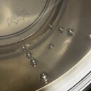 Detailní pohled na vnitřek kovového obruče bubnu s řadou náběhů pro ladicí šrouby. Na průhledné membráně je vidět logo s nápisem REMO v rámci stylizo…
